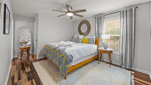 A bedroom with a wooden bed, a ceiling fan, a round mirror, and a window with blinds. - Asheville NC Cottage~Close to shopping and dining~ (Asheville)