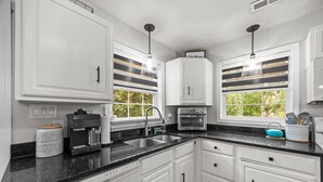 A kitchen with white cabinets, a black countertop, a coffee maker, a toaster, and a window with a view of greenery. - Asheville NC Cottage~Close to shopping and dining~ (Asheville)