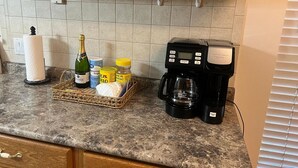 Eine Küchenarbeitsplatte mit Kaffeemaschine, einer Champagnerflasche, einem Brötchenkorb und einem Marmeladenglas.