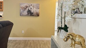 Ein Wohnzimmer mit einem an der Wand hängenden Gemälde eines Pferdes, einer weißen Orchideenpflanze und einer goldenen Elefantenstatue auf einem strukturierten Tisch.
