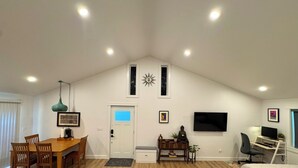 Amplia sala de estar con techos altos, comedor con mobiliario de madera, televisor montado en la pared y una acogedora zona de camas.