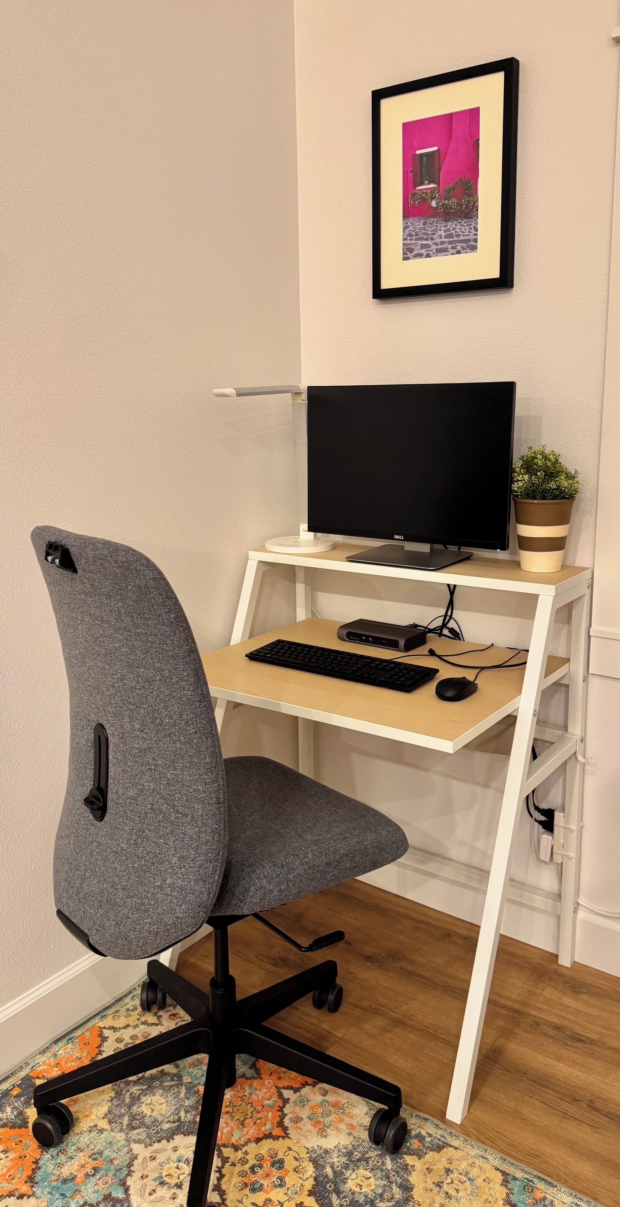A workspace with a grey office chair, a desk with a computer monitor, keyboard, and mouse, and a framed picture on the wall.