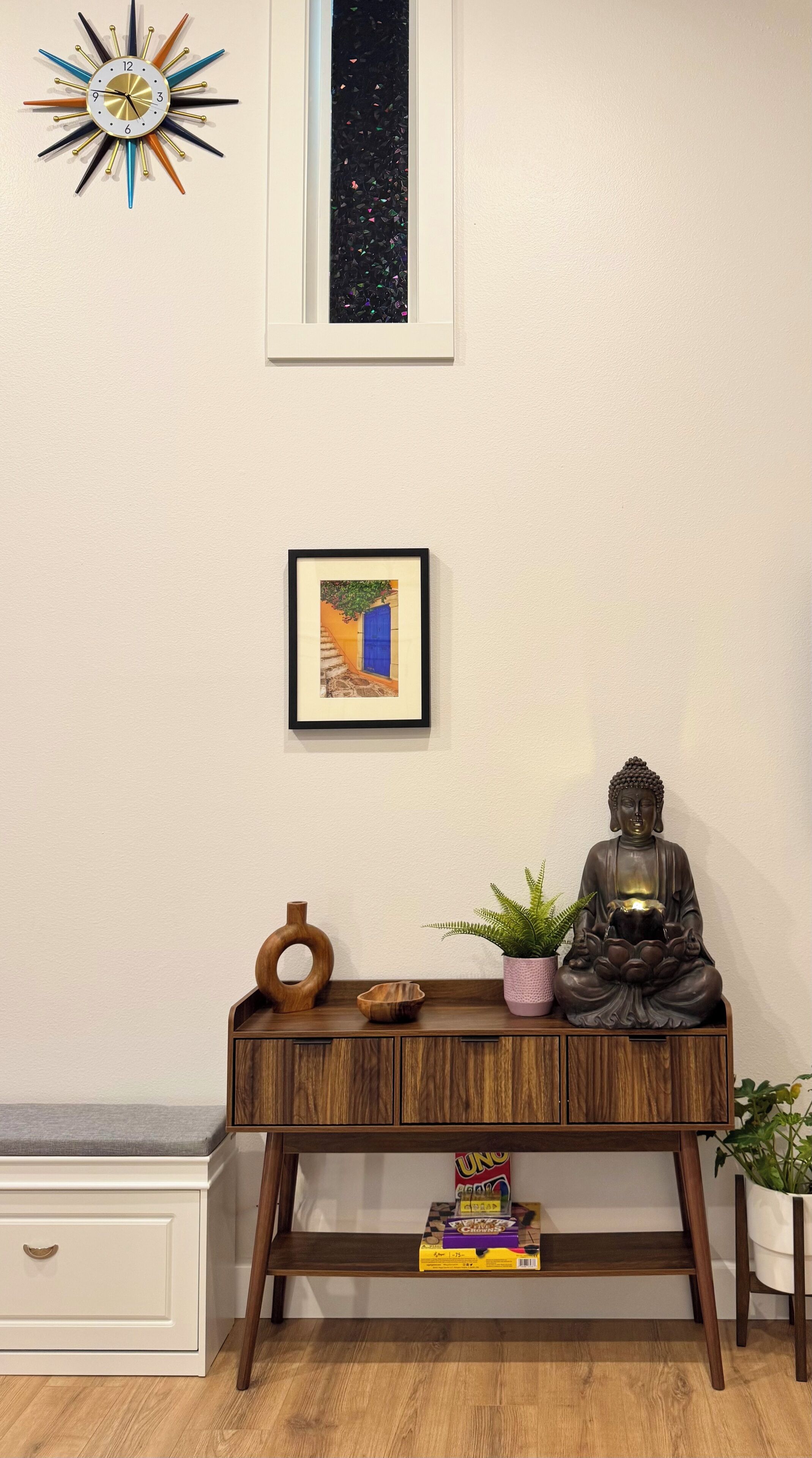 A room with a wooden console table, a Buddha statue, a framed picture, and a starburst clock on the wall.
