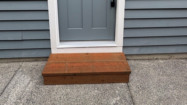 Puerta frontal gris con un panel de vidrio, un timbre negro y un pequeño escalón de madera.