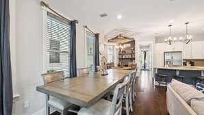 Ein geräumiger Essbereich mit einem langen Holztisch und weißen Stühlen, eine Küche mit weißen Schränken und einer Bar, sowie eine gemütliche Sitzecke mit einer Couch und Sesseln.