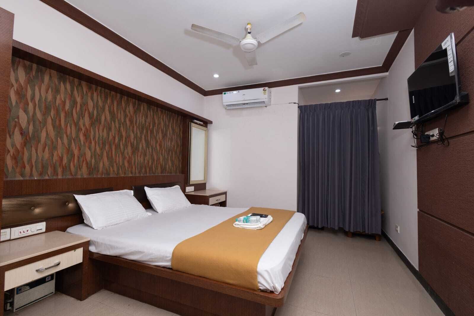 A hotel room with a bed, bedside tables, a TV, an air conditioning unit, and a ceiling fan.