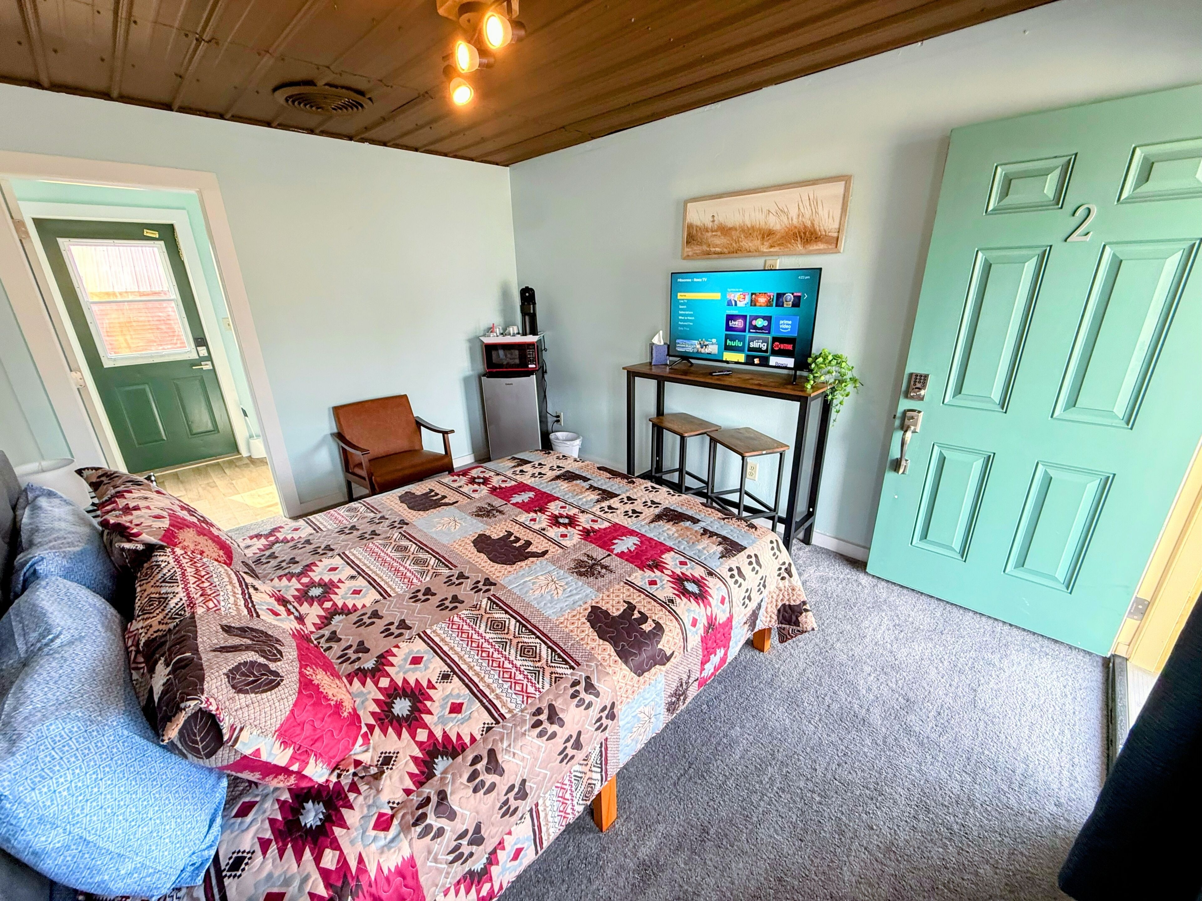 A bedroom with a bed, a chair, a desk with a computer, a TV, and a green door.