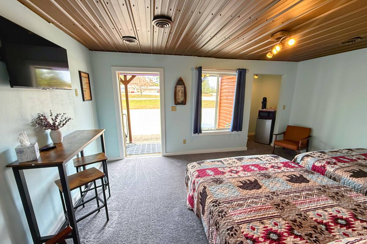 A room with a wooden ceiling, a bed with a patterned blanket, a desk with a vase and a remote control, a chair, and a door leading outside.