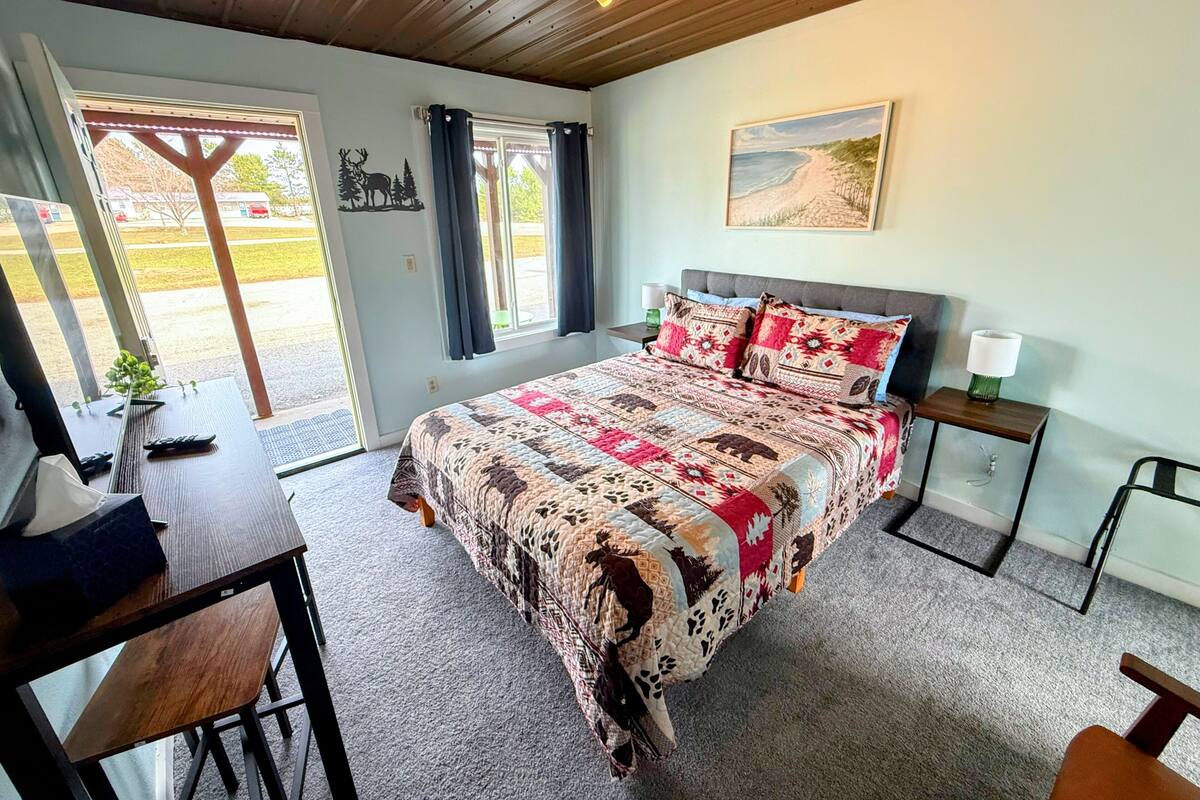 A bedroom with a large bed, a desk, a chair, a nightstand, and a window with curtains.