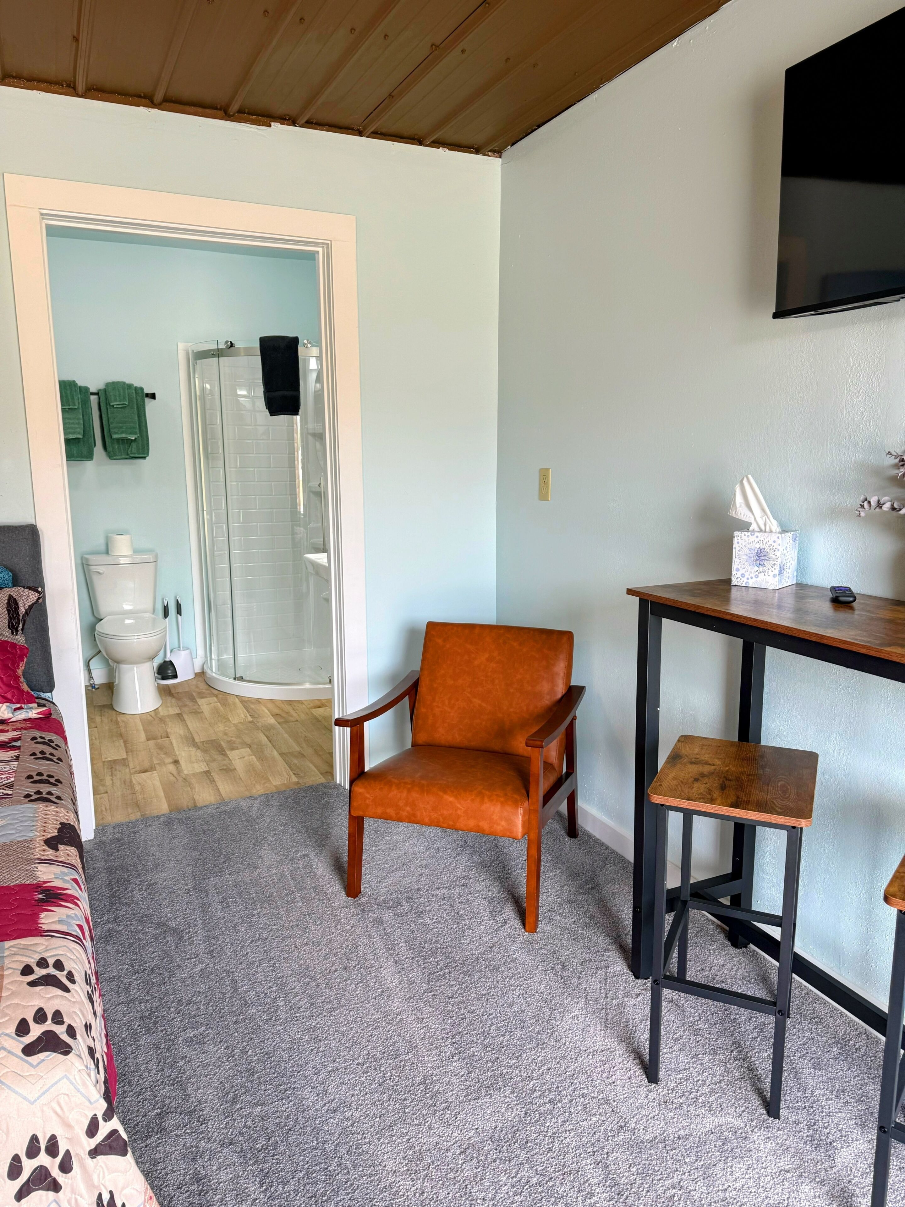 Une pièce avec un plafond en bois, une moquette grise, une chaise en bois, une petite table en bois, un téléviseur fixé au mur et une porte donnant sur une salle de bain équipée de toilettes et d’une douche.