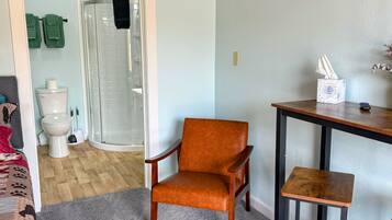 Une pièce avec un plafond en bois, une moquette grise, une chaise en bois, une petite table en bois, un téléviseur fixé au mur et une porte donnant sur une salle de bain équipée de toilettes et d’une douche.