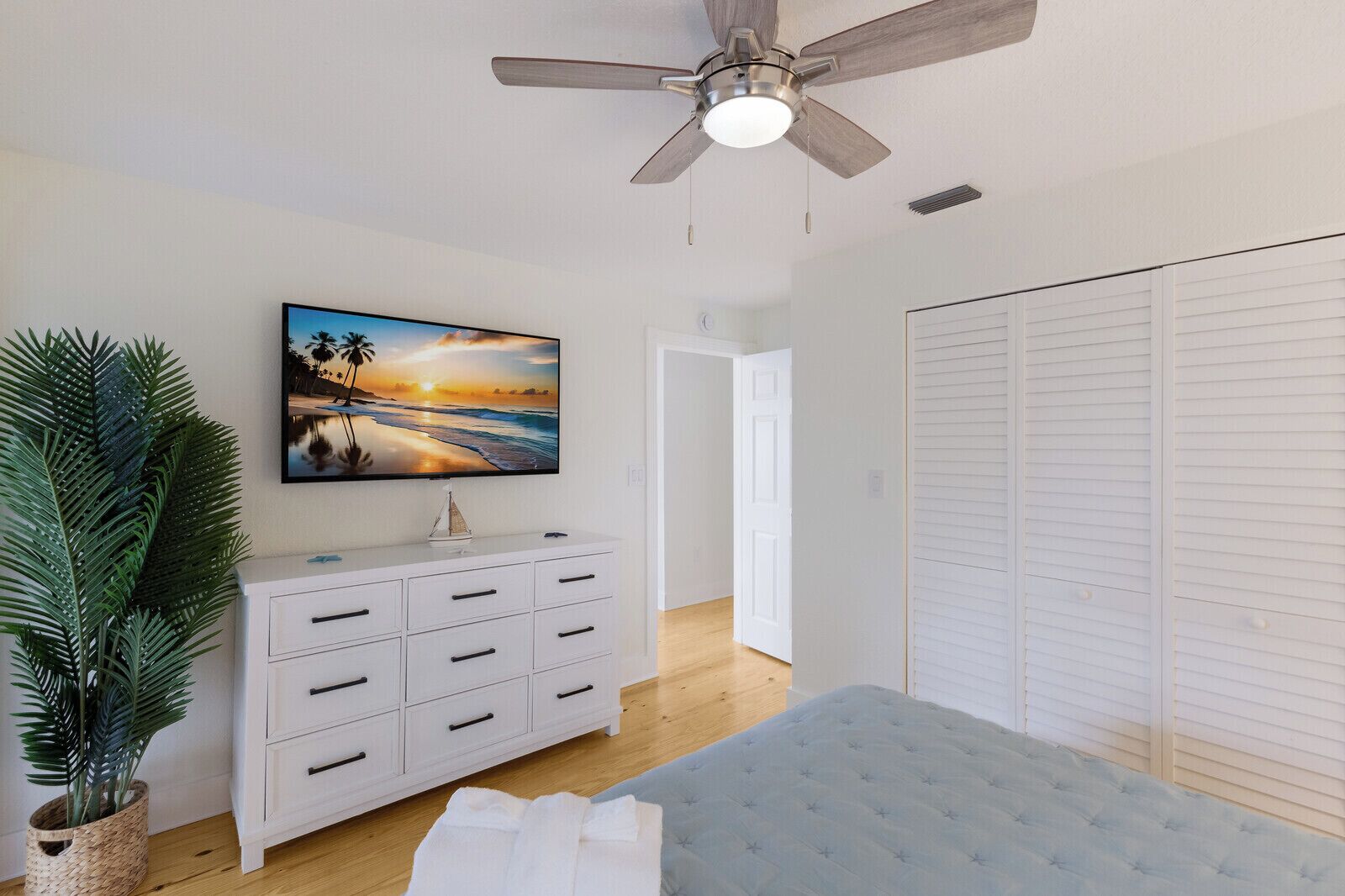Ein Schlafzimmer mit einem Bett, einer weißen Kommode, einem Deckenventilator, einem Fernseher und einer großen Pflanze.