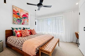 A bedroom with a bed, a bench, a ceiling fan, a window with curtains, and a painting on the wall.
