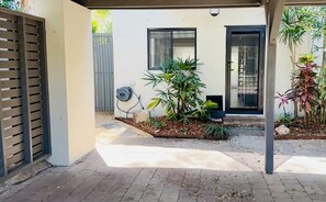 A covered patio area with a tiled floor, a small garden with various plants, a door, and a window.