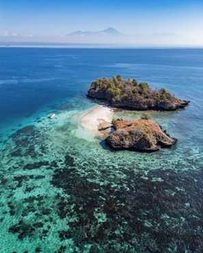 An aerial view of a coastal area with clear turquoise waters, a sandy beach, and rocky outcrops with sparse vegetation. - Satu Hati Hotel and Restaurant (Jerowaru)