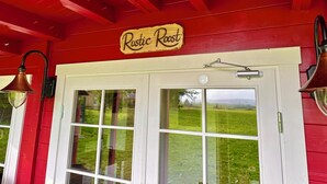 A red building with a sign reading "Rustic Roost" above a white door. - Rustic Roost: Countryside, Warm Cabin w/ Countryside Views & King Bed (Maybole)