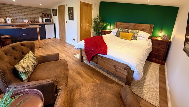 A bedroom with a bed, a wooden headboard, a nightstand, a bedside table, a red throw blanket, and a green wall. - Rustic Roost: Countryside, Warm Cabin w/ Countryside Views & King Bed (Maybole)