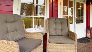 Two wicker chairs with grey cushions on a porch with red siding and white doors. - Rustic Roost: Countryside, Warm Cabin w/ Countryside Views & King Bed (Maybole)