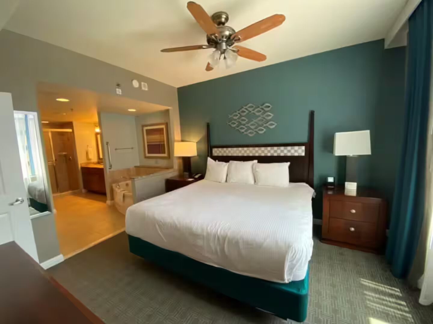 A hotel room with a large bed, a ceiling fan, a nightstand with a lamp, and a bathroom visible through an open door.