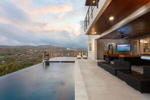 Un área de piscina moderna al aire libre con vista al paisaje, un bar con asientos y un televisor.