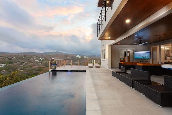 A modern outdoor pool area with a view of the landscape, a bar with seating, and a television.