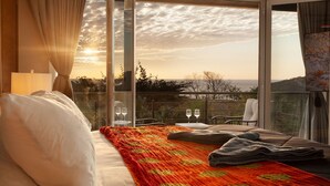 A hotel room with a bed, a view of the sunset, and a balcony with a table and chairs.