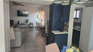 Eine moderne Küche mit dunkelblauen Schränken, einer weißen Arbeitsplatte und einem eingebauten Backofen. Es gibt einen Essbereich mit einem weißen Tisch und Stühlen, und ein graues Sofa im Wohnzimmer.