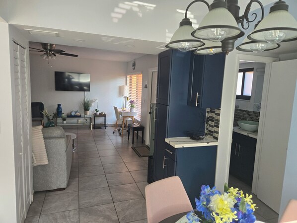 Eine moderne Küche mit dunkelblauen Schränken, einer weißen Arbeitsplatte und einem eingebauten Backofen. Es gibt einen Essbereich mit einem weißen Tisch und Stühlen, und ein graues Sofa im Wohnzimmer.