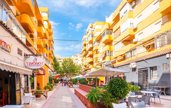 A street with yellow buildings, outdoor seating, and potted plants.