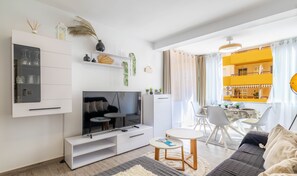 A modern living room with a flat-screen TV, white cabinetry, a dining area with a table and chairs, and a view of a yellow building through the window.