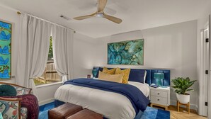 Une chambre à coucher avec un lit, un ventilateur de plafond, une table de chevet, une plante en pot et des œuvres d’art accrochées aux murs.