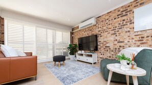 A living room with a brick wall, a flat-screen TV, a sofa, a coffee table, and a window with blinds. - Nelson Bay Family Retreat (Nelson Bay)