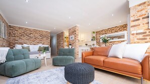 A modern living room with a brick wall, two green sofas, an orange leather sofa, and a round ottoman. - Nelson Bay Family Retreat (Nelson Bay)