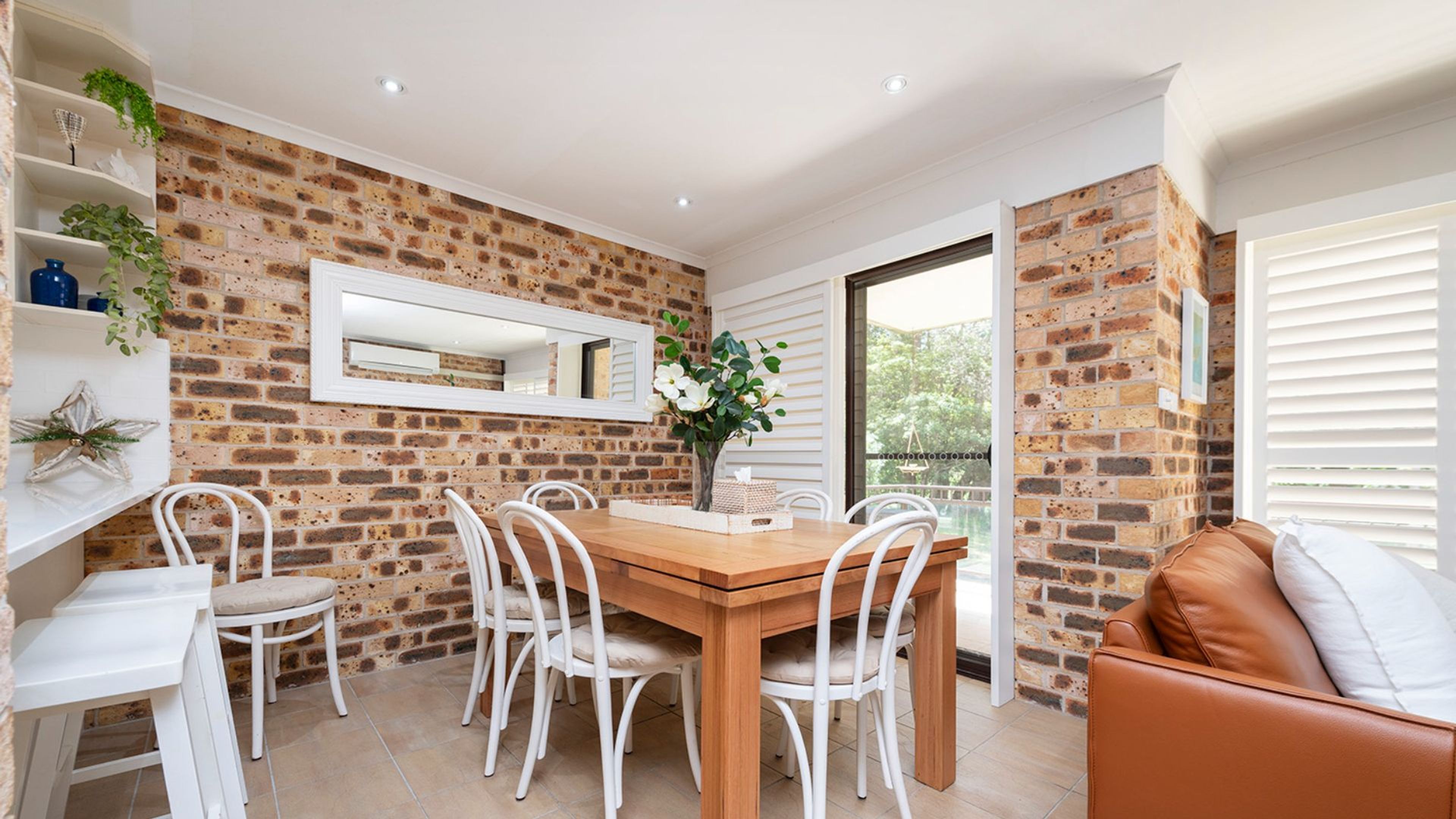 Un comedor con pared de ladrillo, sillas blancas, mesa de madera y sofá de cuero marrón.