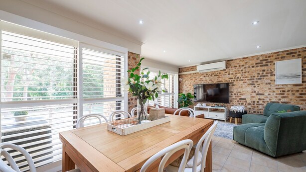 A modern living room with a brick wall, a flat-screen TV, a wooden dining table, and a green sofa. - Nelson Bay Family Retreat (Nelson Bay)
