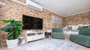 A living room with a brick wall, a flat-screen TV, a green sofa, a white console table, and a potted plant.