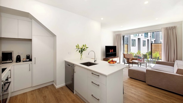 Een moderne keuken met witte kasten, een inbouwvaatwasser en een centraal eiland met een spoelbak. Er is een zithoek met een bank en een televisie op de achtergrond.