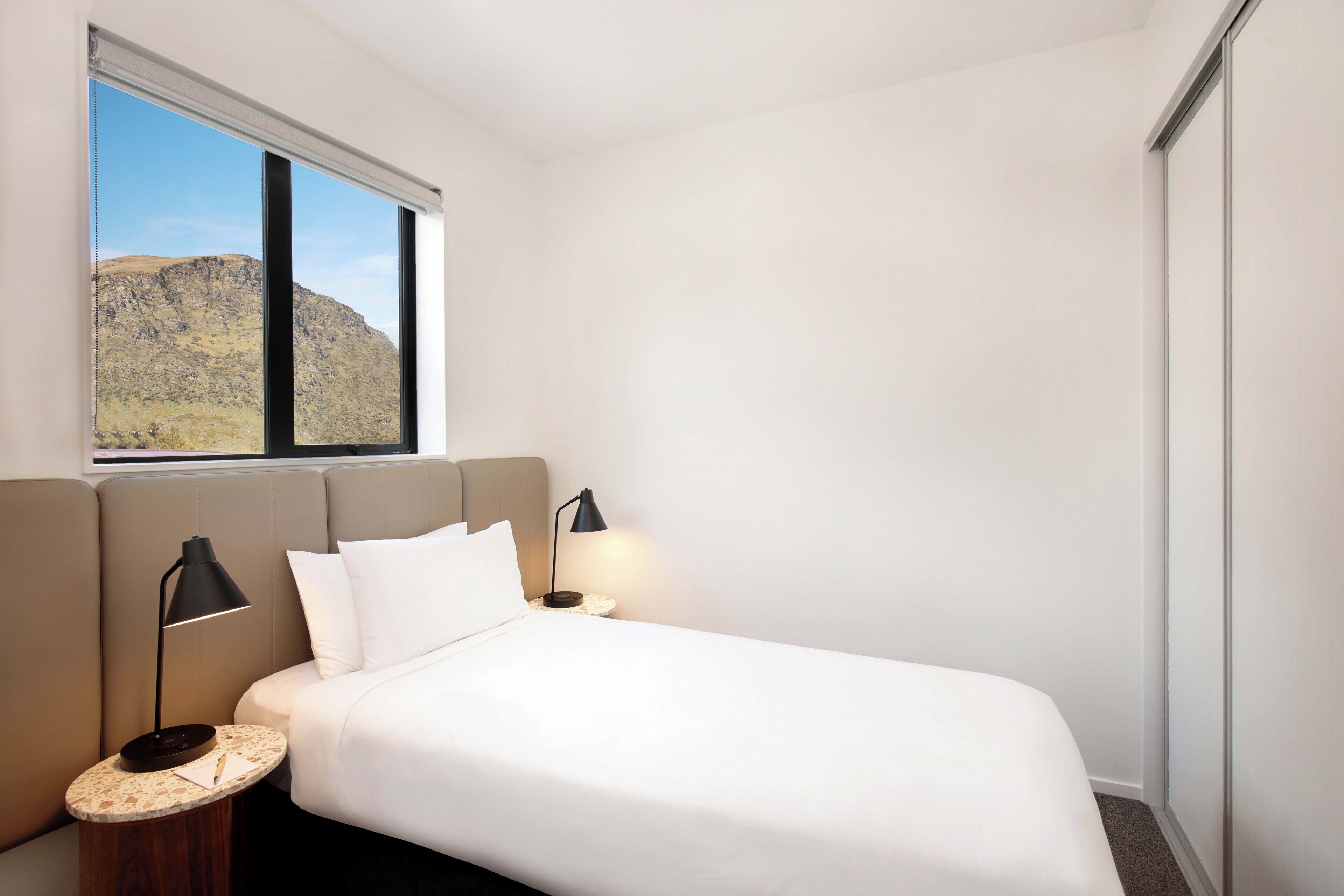 A modern hotel room with a large bed, two bedside tables, a window with a mountain view, and a neutral color scheme.