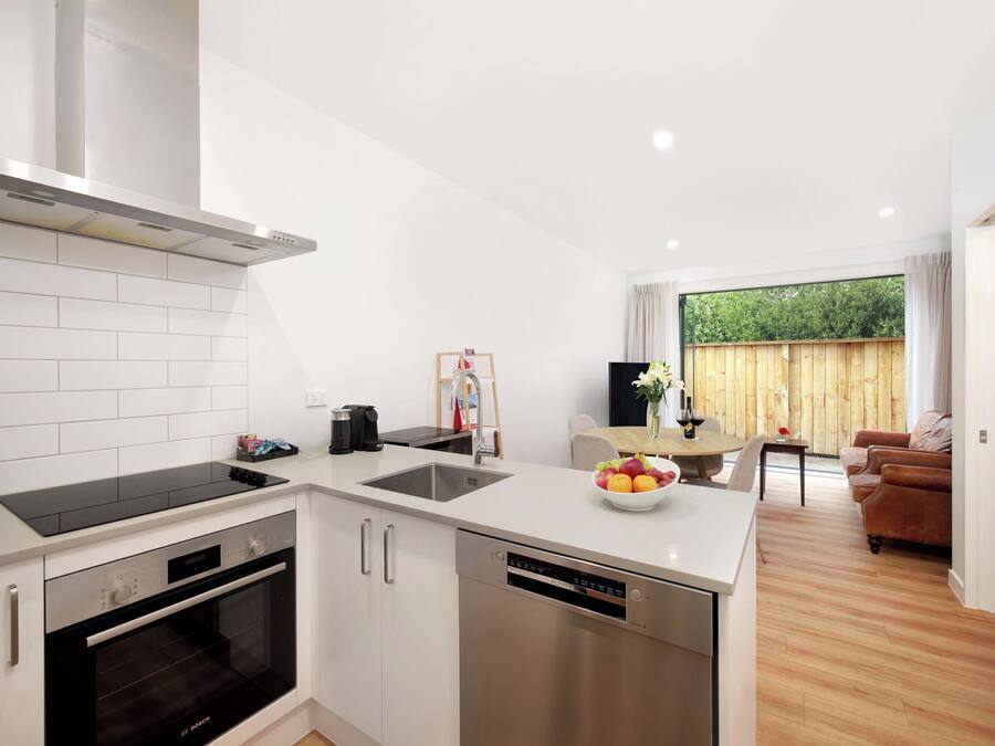 A modern kitchen with white cabinets, a dishwasher, and a built-in oven. There is a dining area with a wooden table and chairs, and a view of a wooden fence outside.