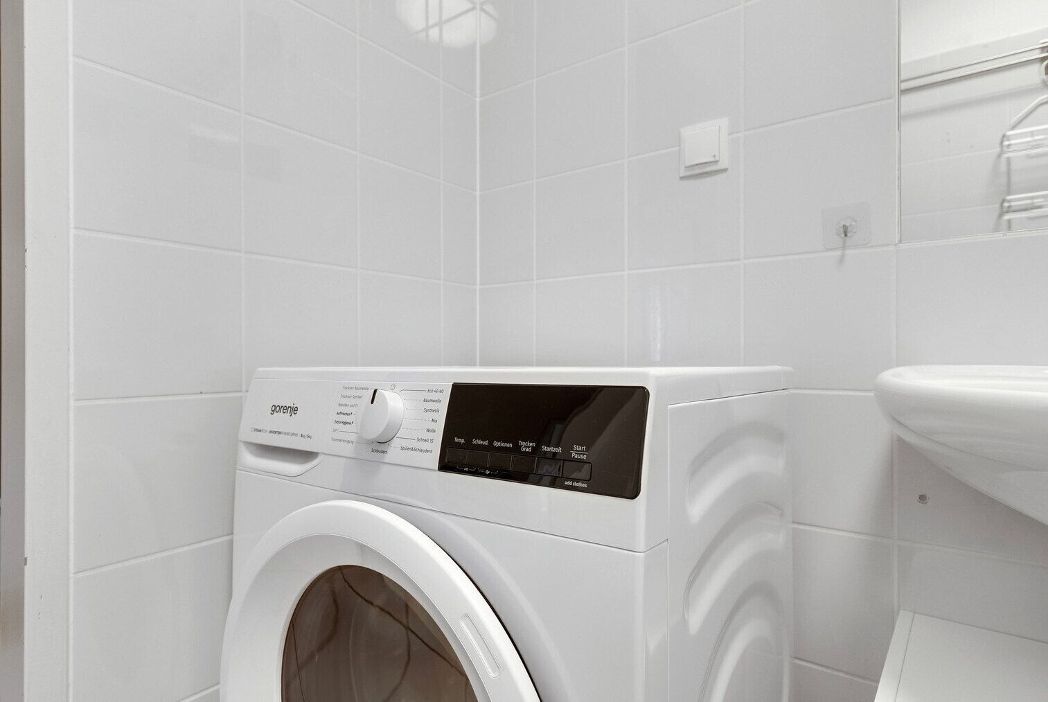 A front-loading washing machine in a bathroom with white tiled walls and a white sink.