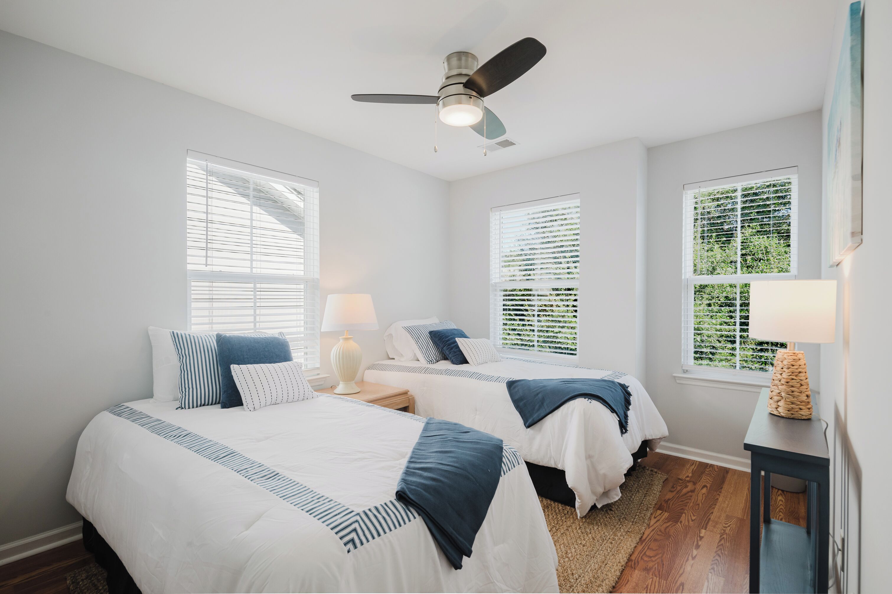 Une chambre avec deux lits, un ventilateur de plafond et de grandes fenêtres équipées de stores.