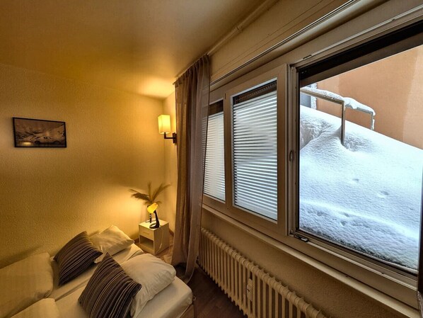 A hotel room with a bed, a nightstand with a lamp, a radiator, and a window with blinds.