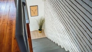 Una escalera con suelo de madera que lleva a una plataforma, una planta en maceta y una pared con líneas verticales.