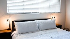 Un dormitorio con cama, ropa de cama blanca, dos lámparas de noche y una ventana con persianas.