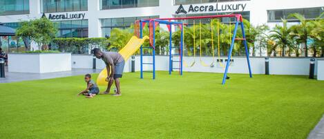 A playground with a yellow slide and a swing set, surrounded by artificial grass, in front of a building with 'Accra Luxury' signage.