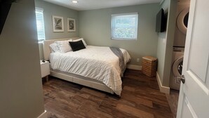 A bedroom with a bed, a nightstand, a wall-mounted TV, a window with blinds, and a washing machine in the corner. - Inland Escape - Centrally Located (Jacksonville)