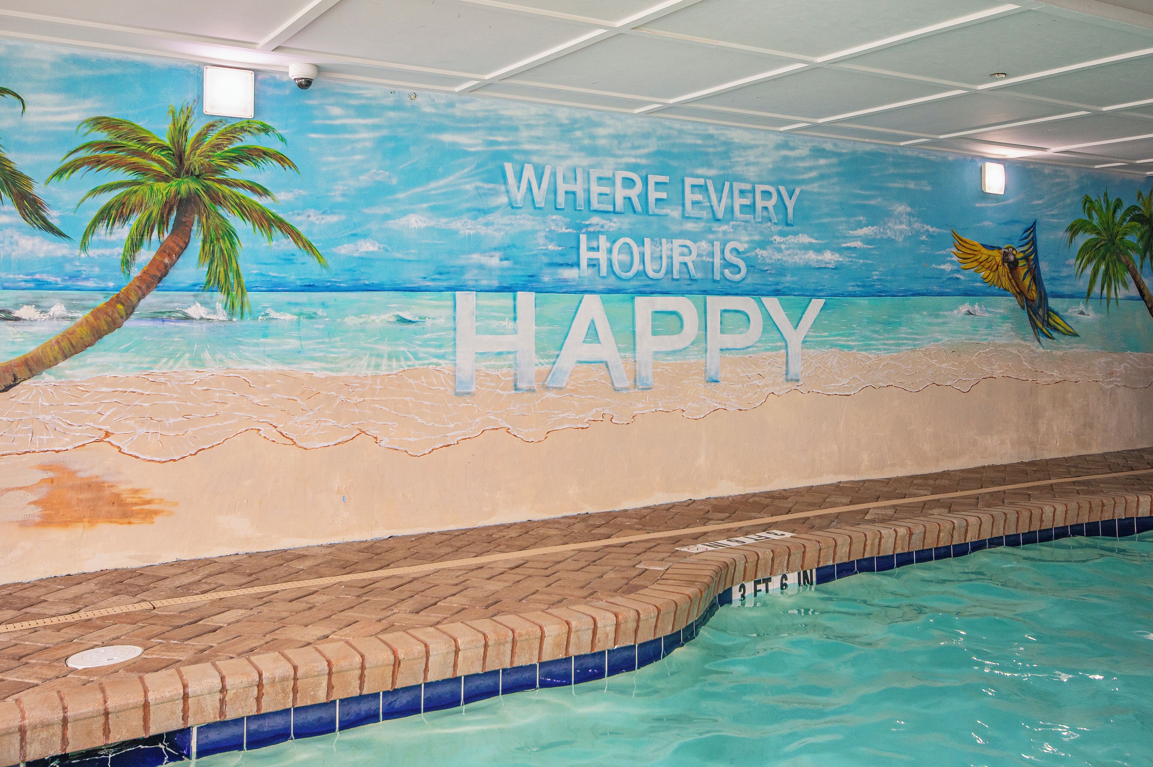 Ein Schwimmbecken mit einem Wandgemälde, das eine Strandlandschaft zeigt, und dem Schriftzug 'WHERE EVERY HOUR IS HAPPY'.