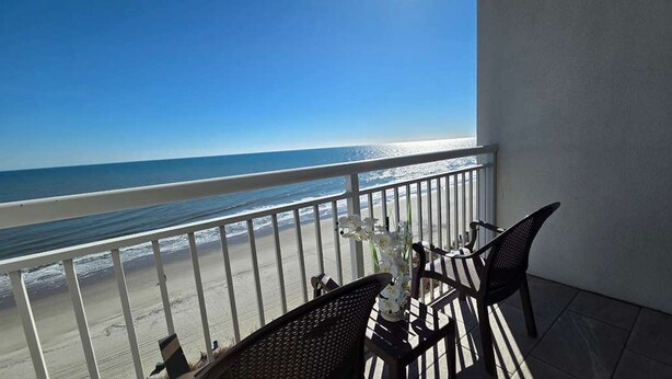 A balcony with a view of the beach and ocean. There are two chairs and a table with a vase of flowers. - Oceanfront One-Bedroom Condo with Stunning Paradise Views (Myrtle Beach)