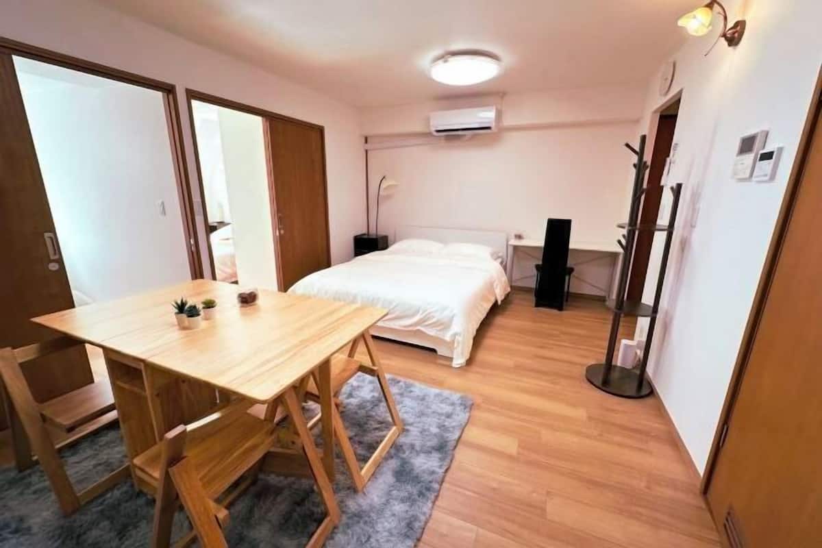 Een kamer met een bed, een houten tafel, een bureau, een grijze vloerkleed en een plafondlamp.