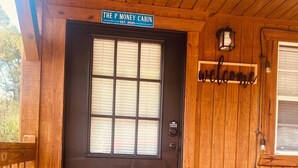 Una cabaña de madera con una puerta marrón oscuro, un letrero de bienvenida encima de la puerta, una silla de metal a la derecha y una terraza de madera con felpudo.